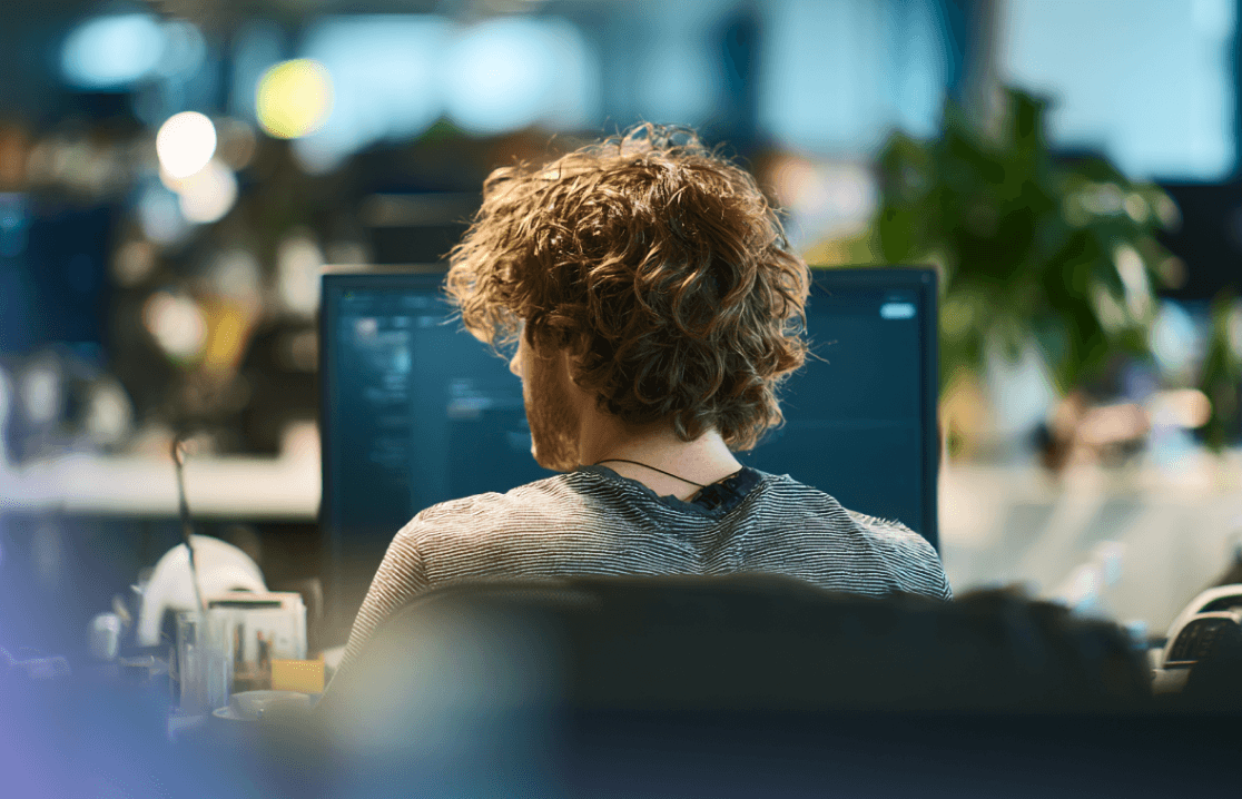 Vue par-dessus l'épaule d'un développeur aux cheveux bouclés travaillant sur un ordinateur dans un bureau moderne.